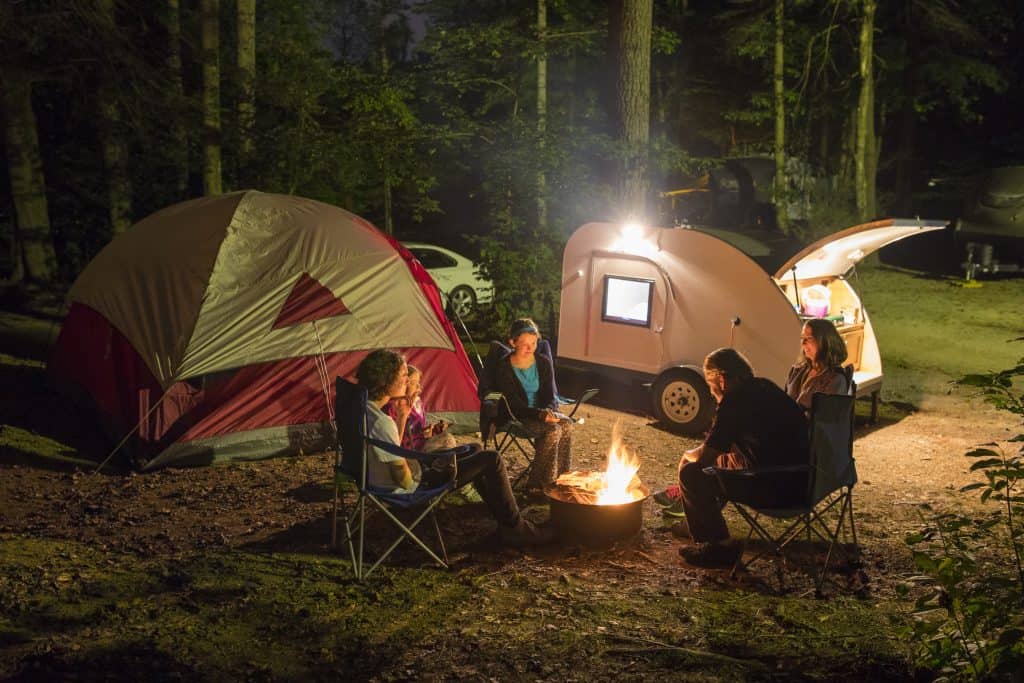 Family enjoys campfire around tent and teardrop trailer.