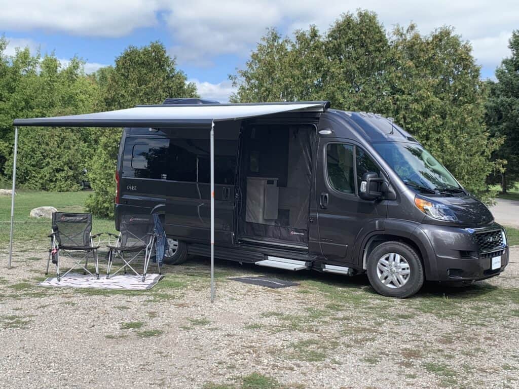 Class B RV at camp with awning and chairs.