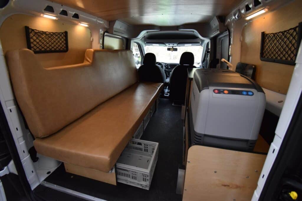 interior of Cascade Campers van conversion featuring a couch, storage bins under couch, a kitchen area with freezer-fridge, and storage pockets on wall
