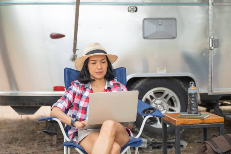 Woman RV camping sits outside her RV using a laptop.