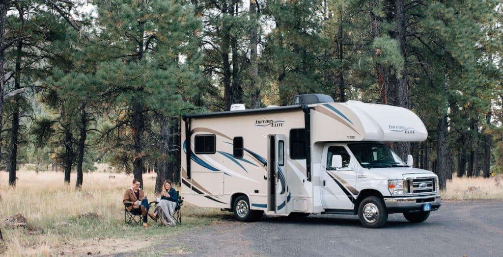 Class C white RV camping in forest  with man and woman sitting in camp chairs - feature image for Where To Buy An RV With Bad Credit