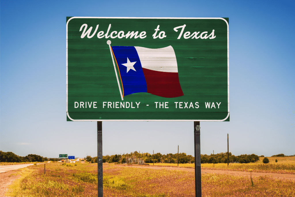 A welcome sign for Texas in the center of a highway. The sign is green with a hand-painted texas flag, and reads “Welcome to Texas. Drive Friendly - The Texas Way”
