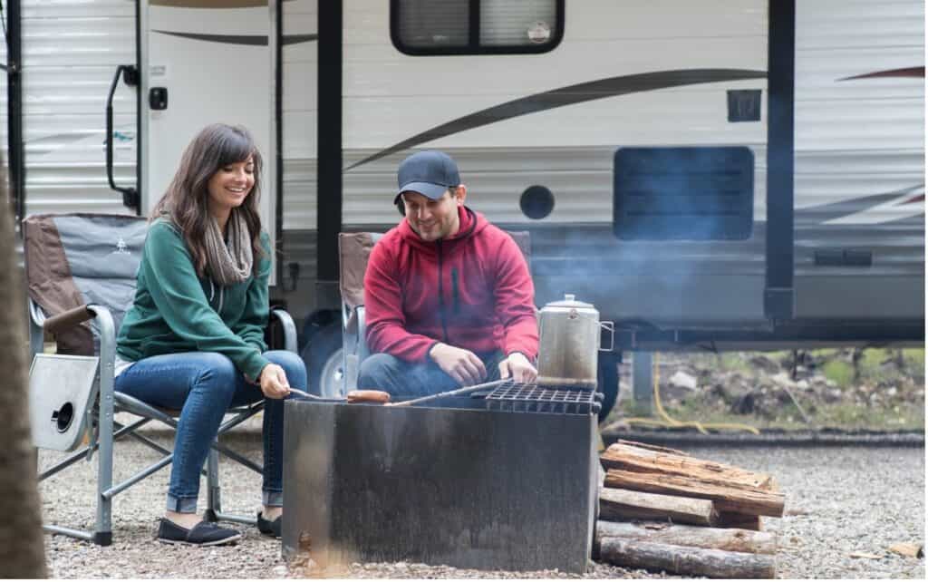 A man and woman roasting hot dogs over a campfire in front of their RV