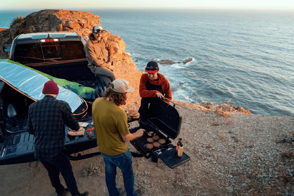 Four young men gathered around a truck with hitch mounted grill cooking burgers beside the ocean - propane grill