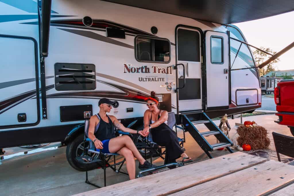 Two women sit in front of a travel trailer.