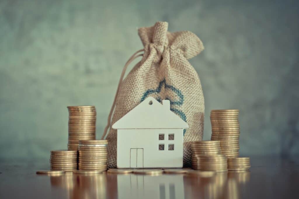 Stacks of coins surrounding a model of a house in front of a gunny sack to imply home equity.