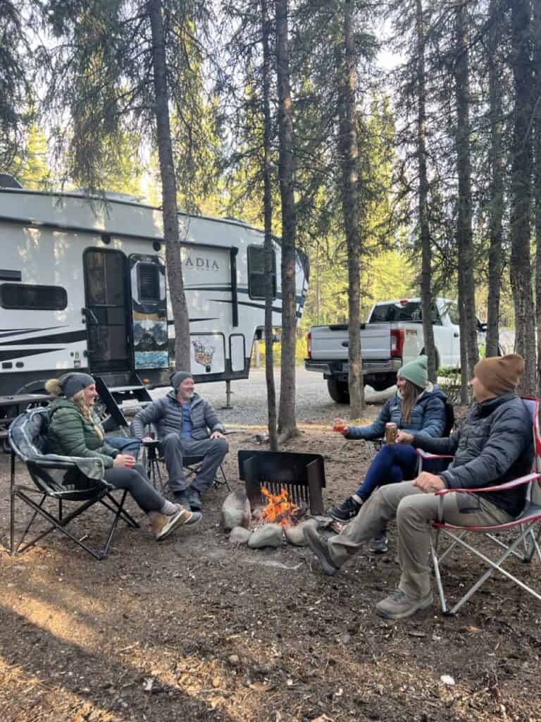 A group of campers around a campfire in fornt of a fifth-wheel and truck.