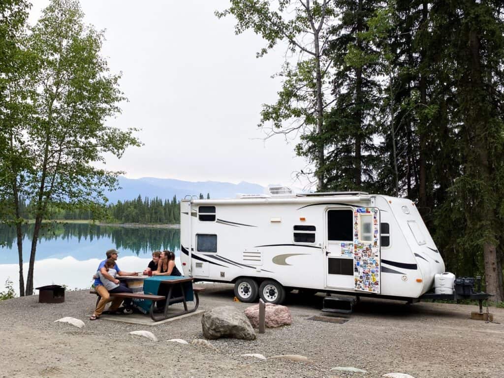 A travel trailer by a lake, with a family eating a meal at a picnic table.